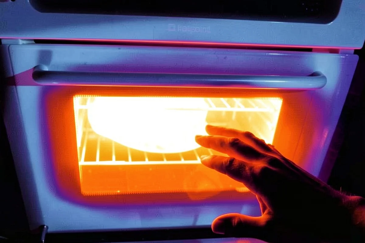 A person's hand reaches toward the glass door of an open oven with a glowing white-hot heating element inside.